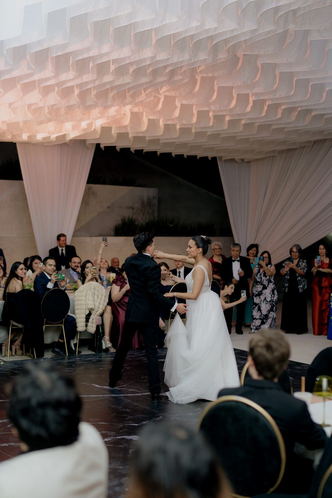 Newlyweds dance on marble floor surrounded by applauding guests under sculptural white ceiling.