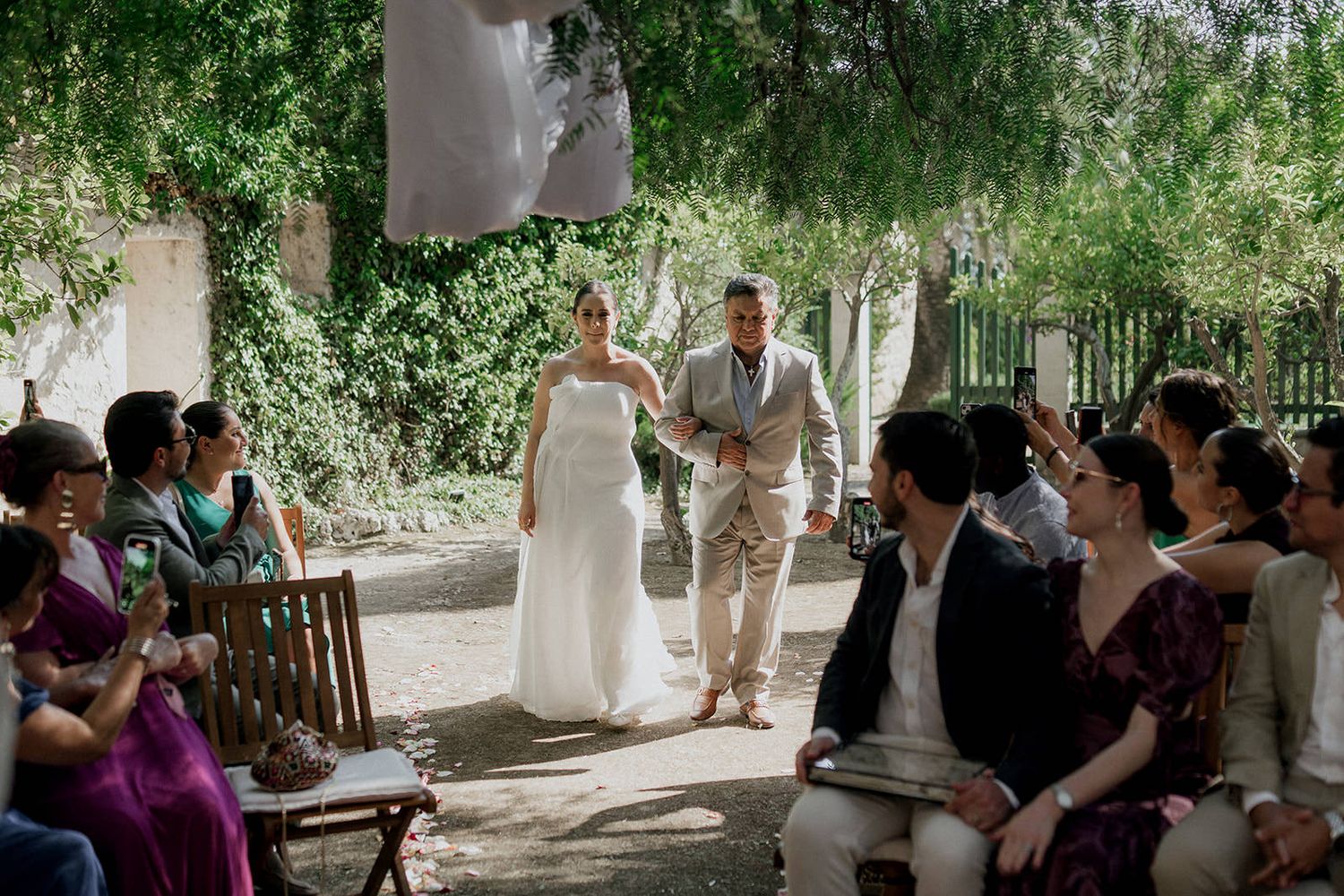 An outdoor wedding ceremony takes place under lush green trees with guests seated on wooden benches.