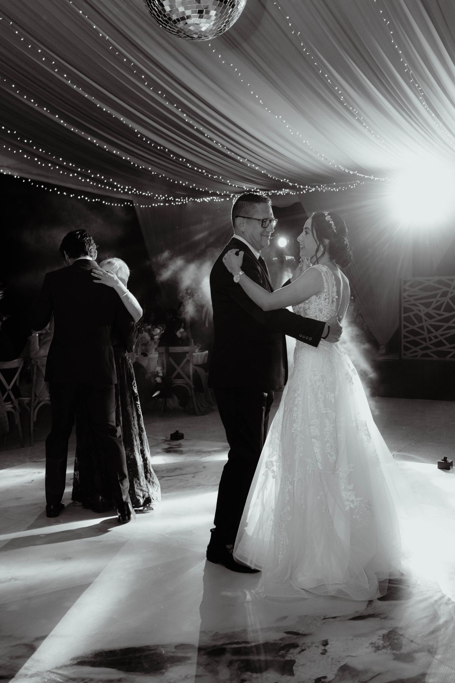 Black and white photo of couples dancing under a disco ball and draped ceiling at a wedding reception.