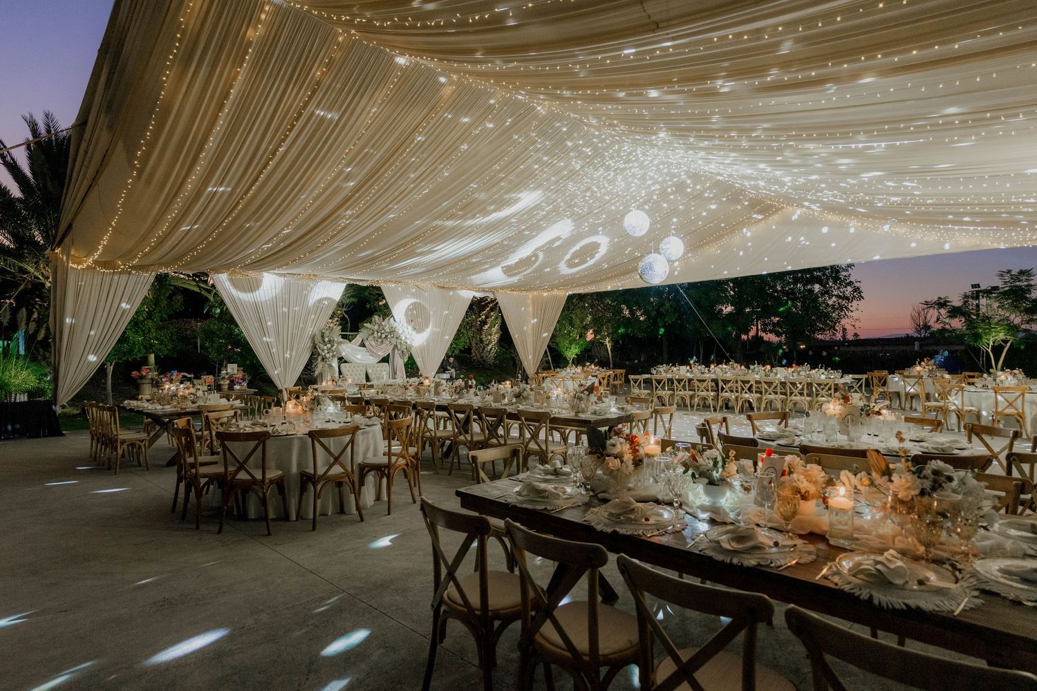 Wedding reception venue features elegant table settings under a draped canopy with twinkling lights.
