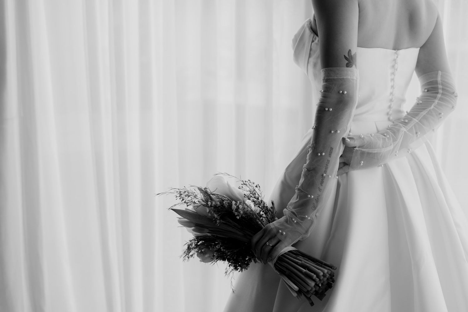 Black and white artistic photo of a bride holding a bouquet against sheer curtains.