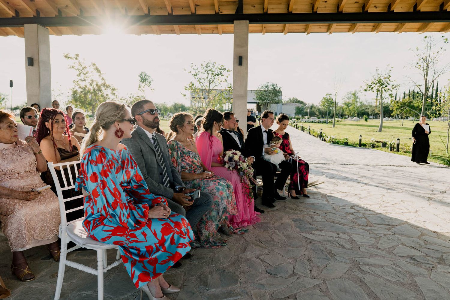 Wedding guests seated in white chairs under covered pavilion during outdoor ceremony.