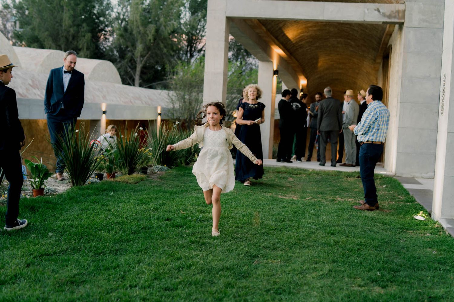 A young flower girl in white dress runs joyfully across grass while wedding guests look on.