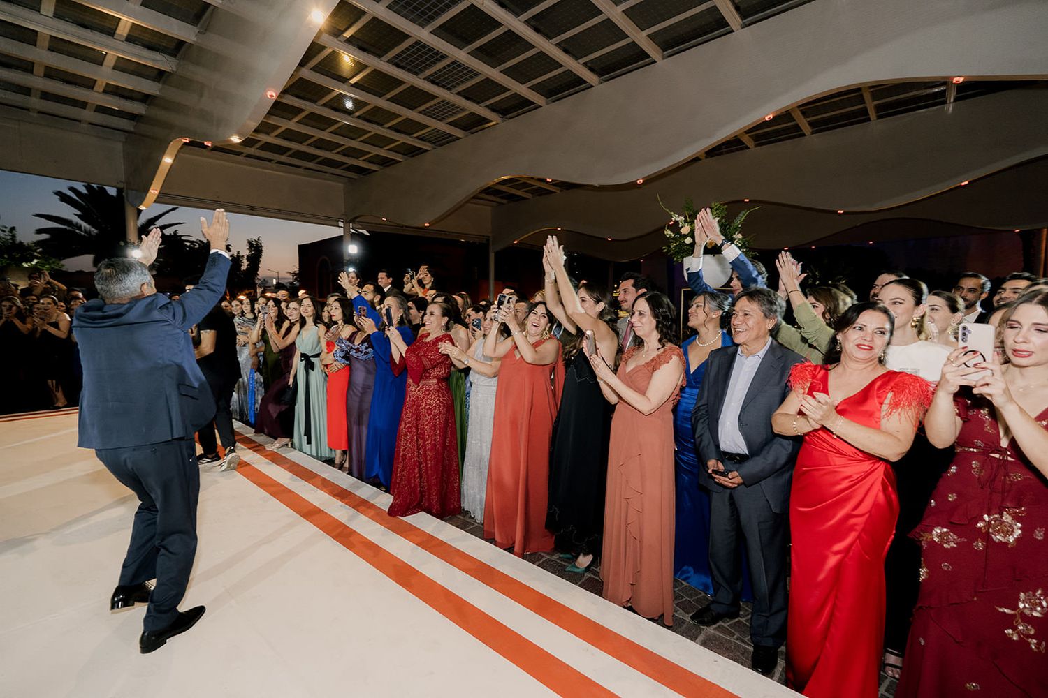 Wedding guests in formal attire celebrate on a white dance floor, raising their hands during a lively reception party.