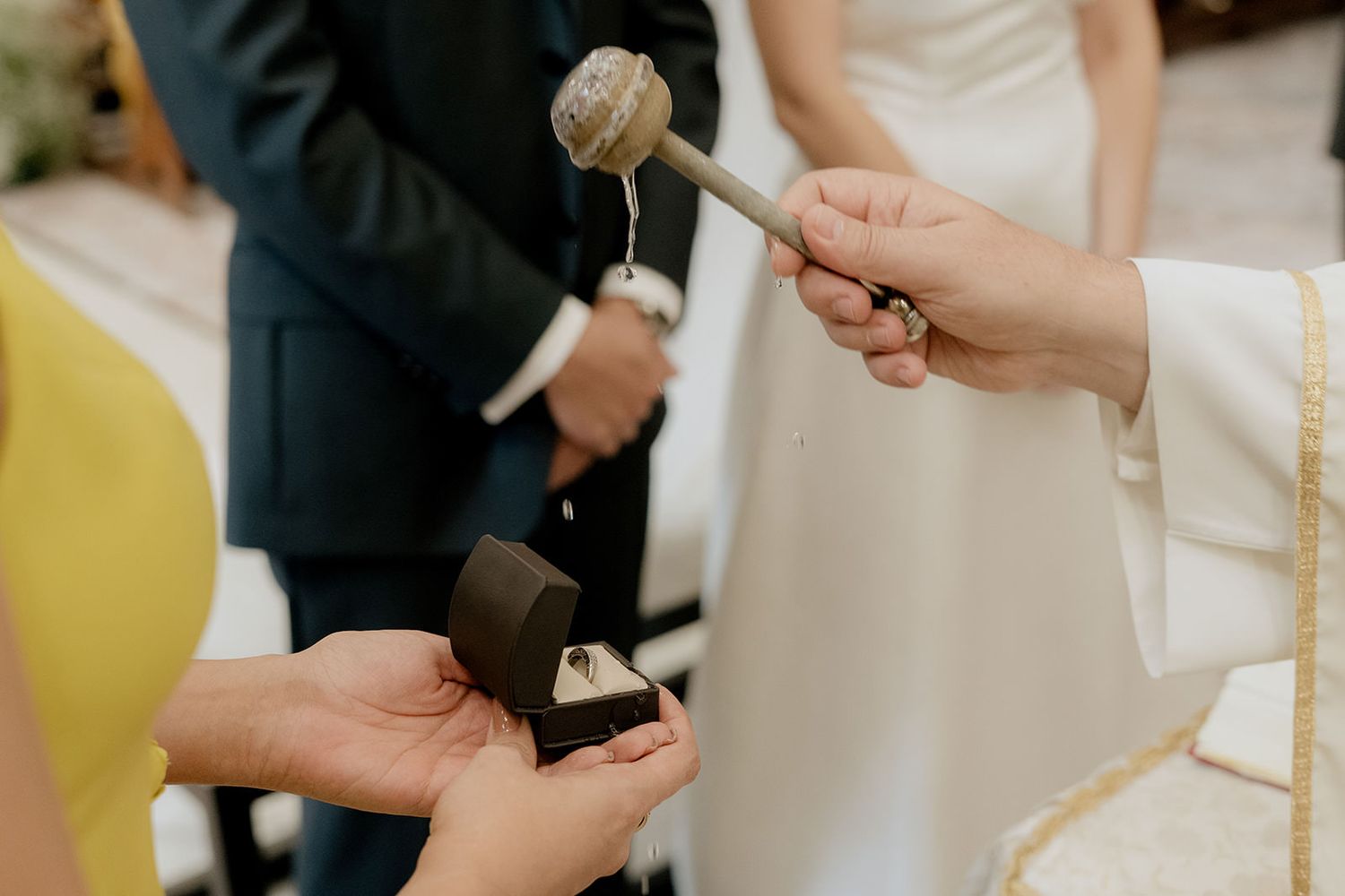 Priest blessing wedding rings in black velvet box with holy water during a traditional church ceremony.
