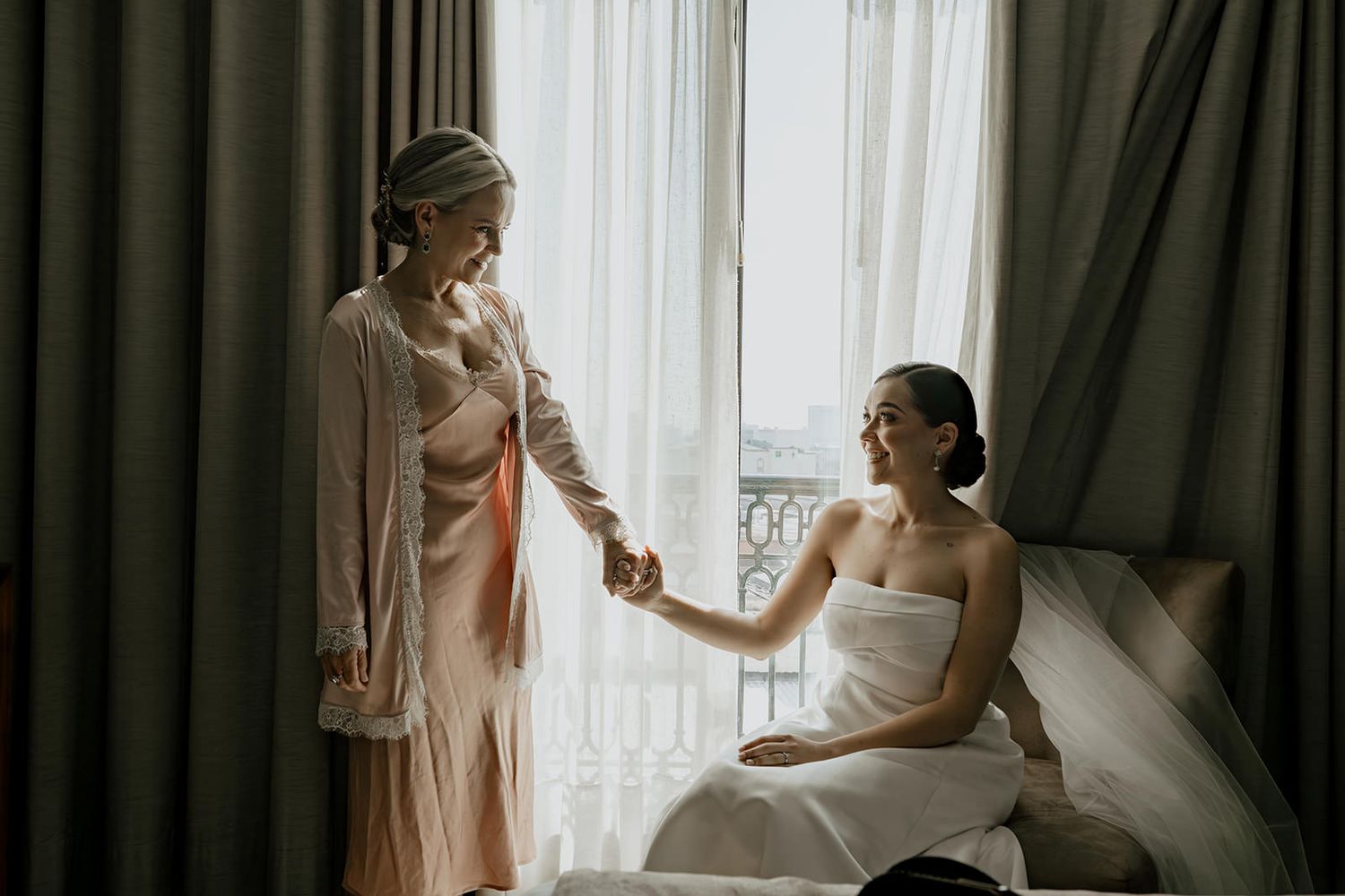 Bride in white strapless gown holds hands with bridesmaid in pink robe by a sunlit window during wedding preparations.