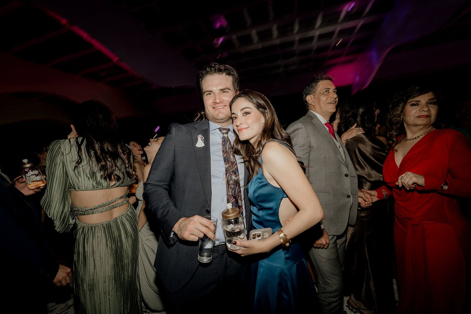 Couple poses together smiling at a wedding reception party with colorful ambient lighting in background.