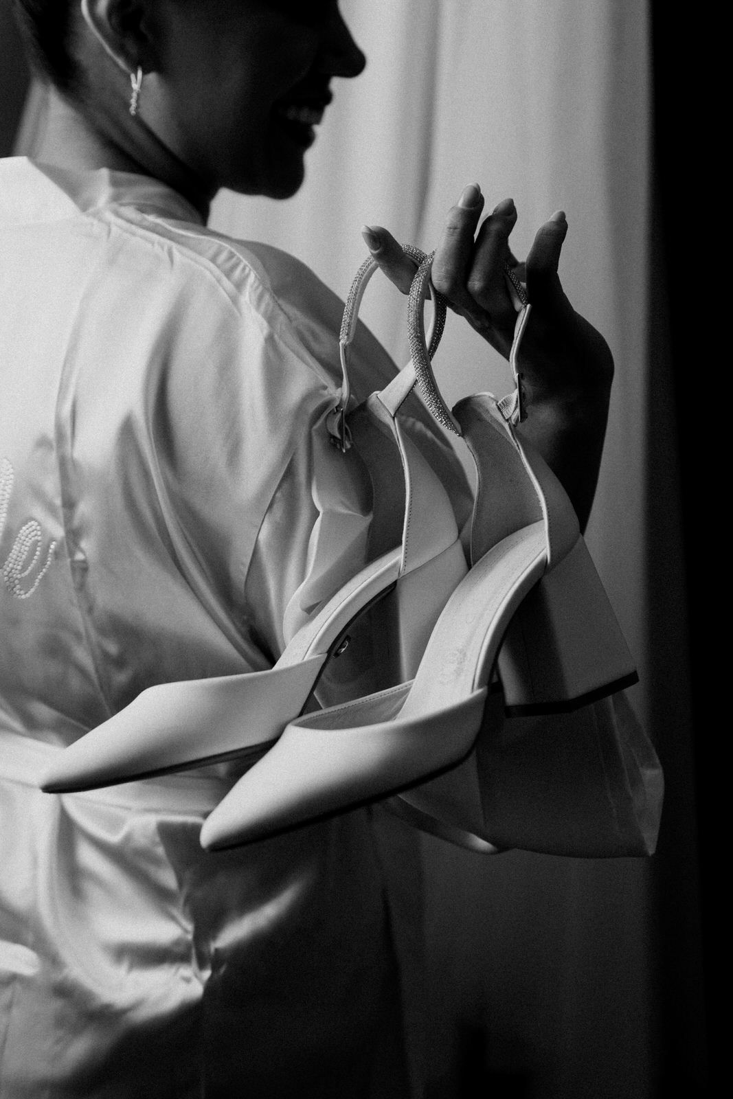 Black and white photo of bride in robe holding white heeled wedding shoes.