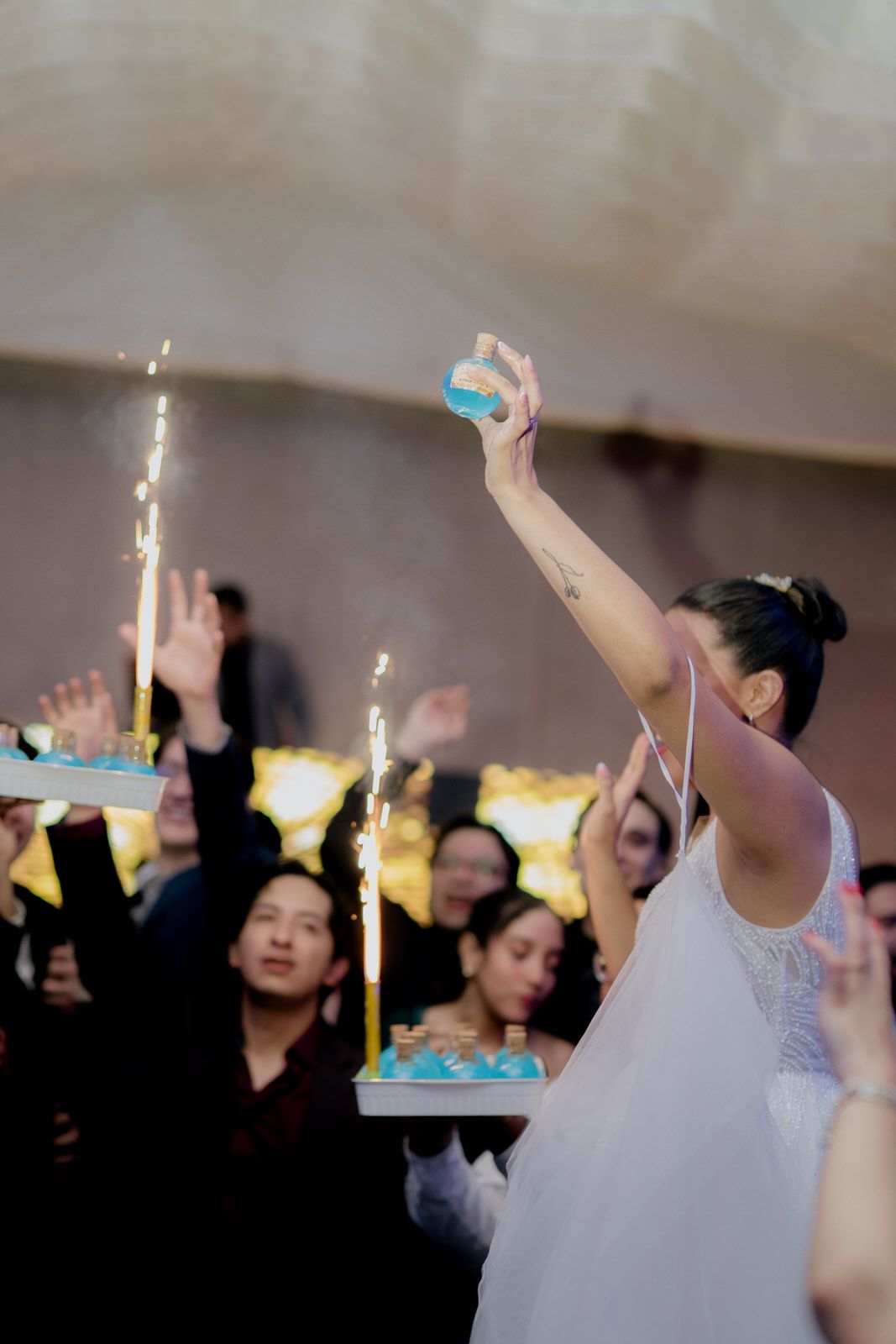 Bride holds blue shot glass aloft with sparklers on trays at a lively wedding party.