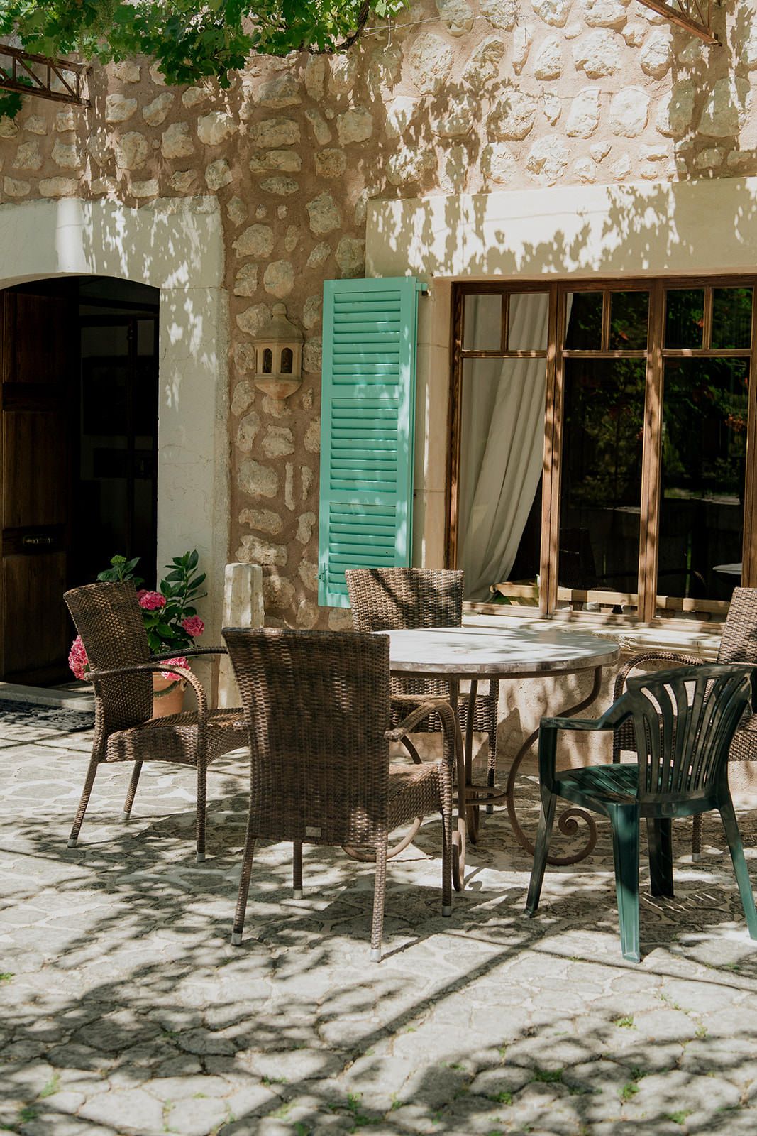 Shaded outdoor patio with stone walls, green shutters, and scattered dining furniture.