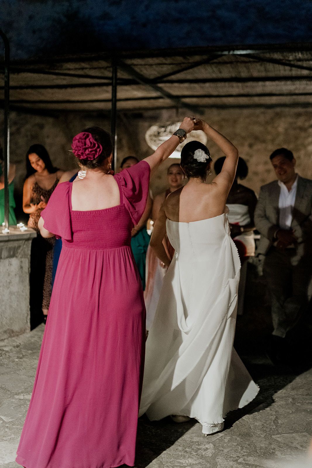 Women dancing at a celebration, one in a pink dress and another in white, with guests in the background.