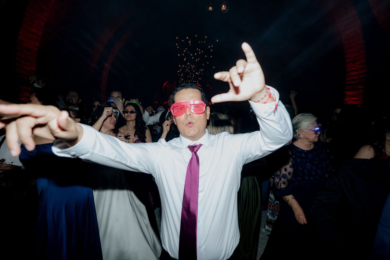 A guest dances enthusiastically wearing pink sunglasses at a wedding reception.