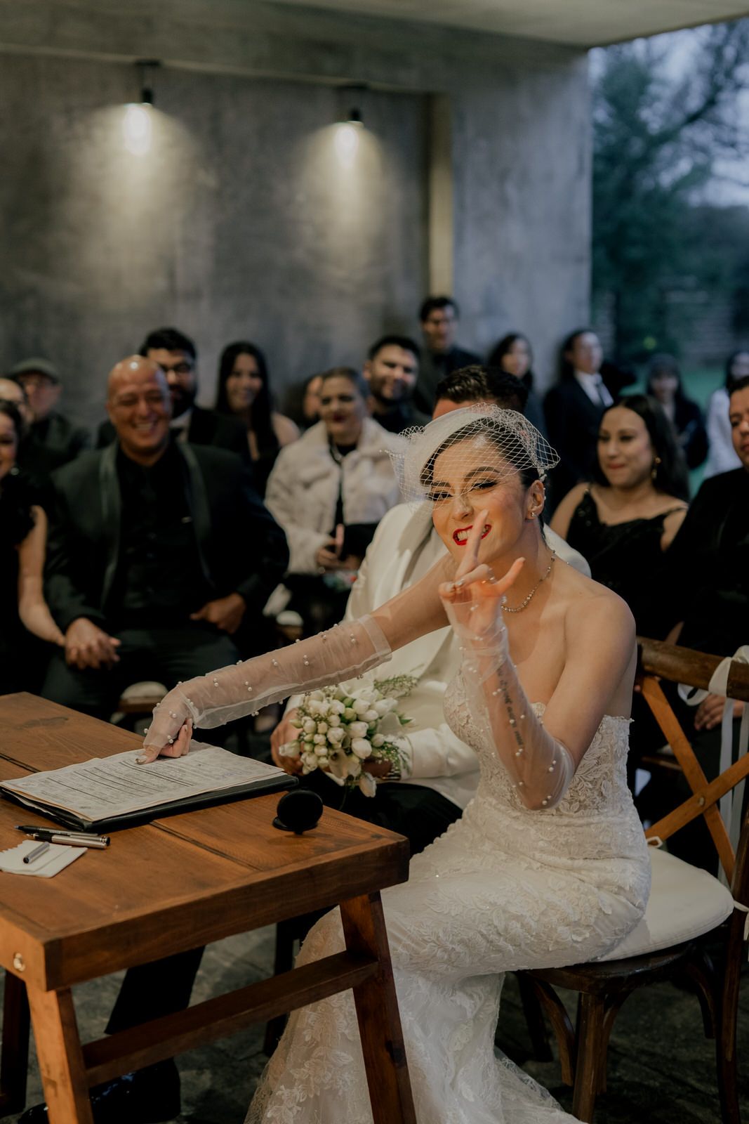 A person in a wedding dress blows a kiss while seated at a wooden table during the ceremony.