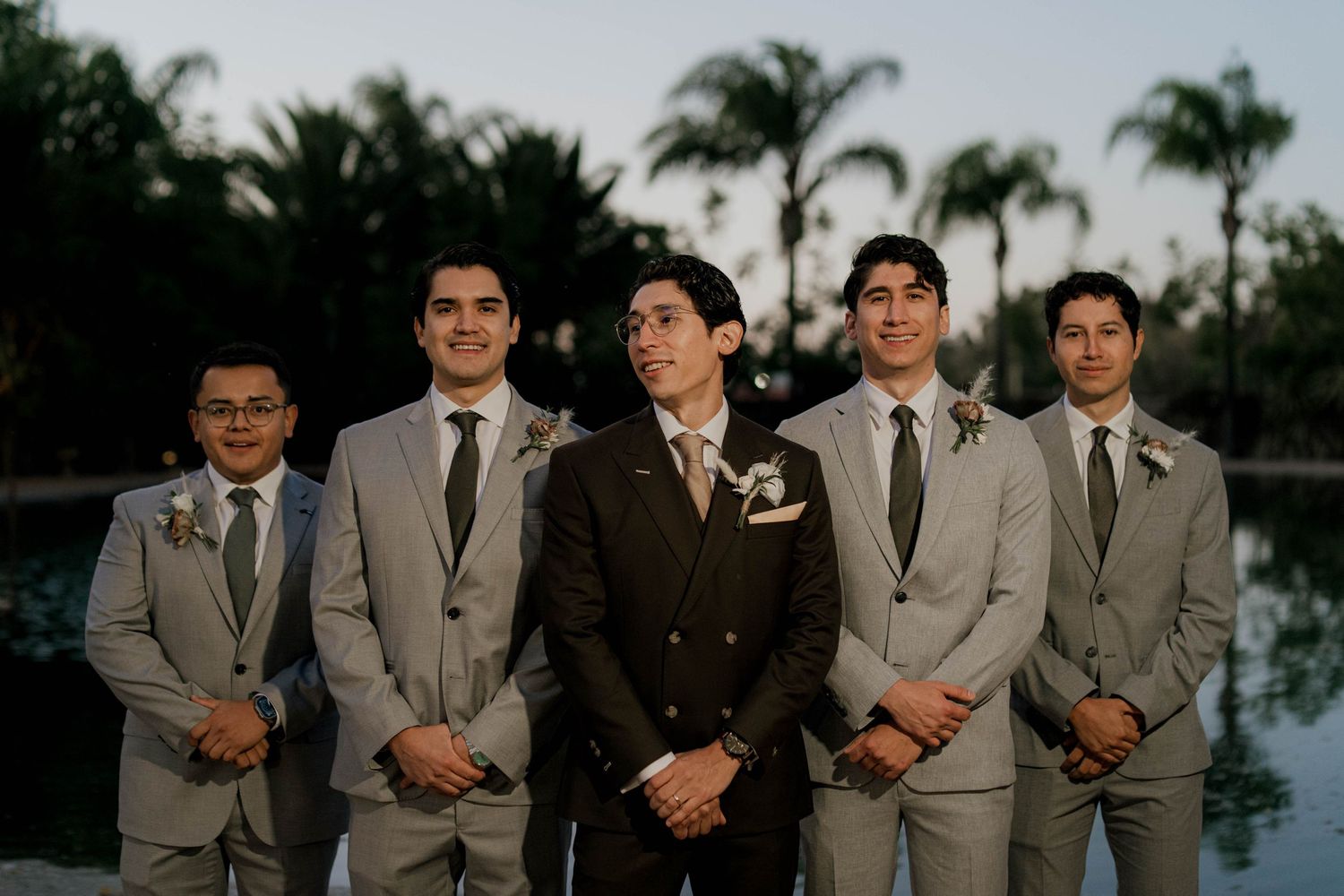 Groomsmen in matching gray suits pose together at sunset near palm trees.