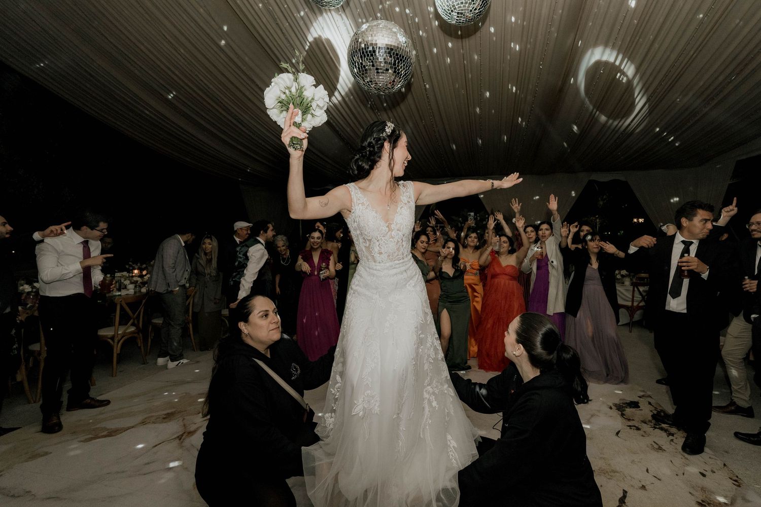 Wedding reception dance floor packed with guests under glowing disco balls and draped ceiling.