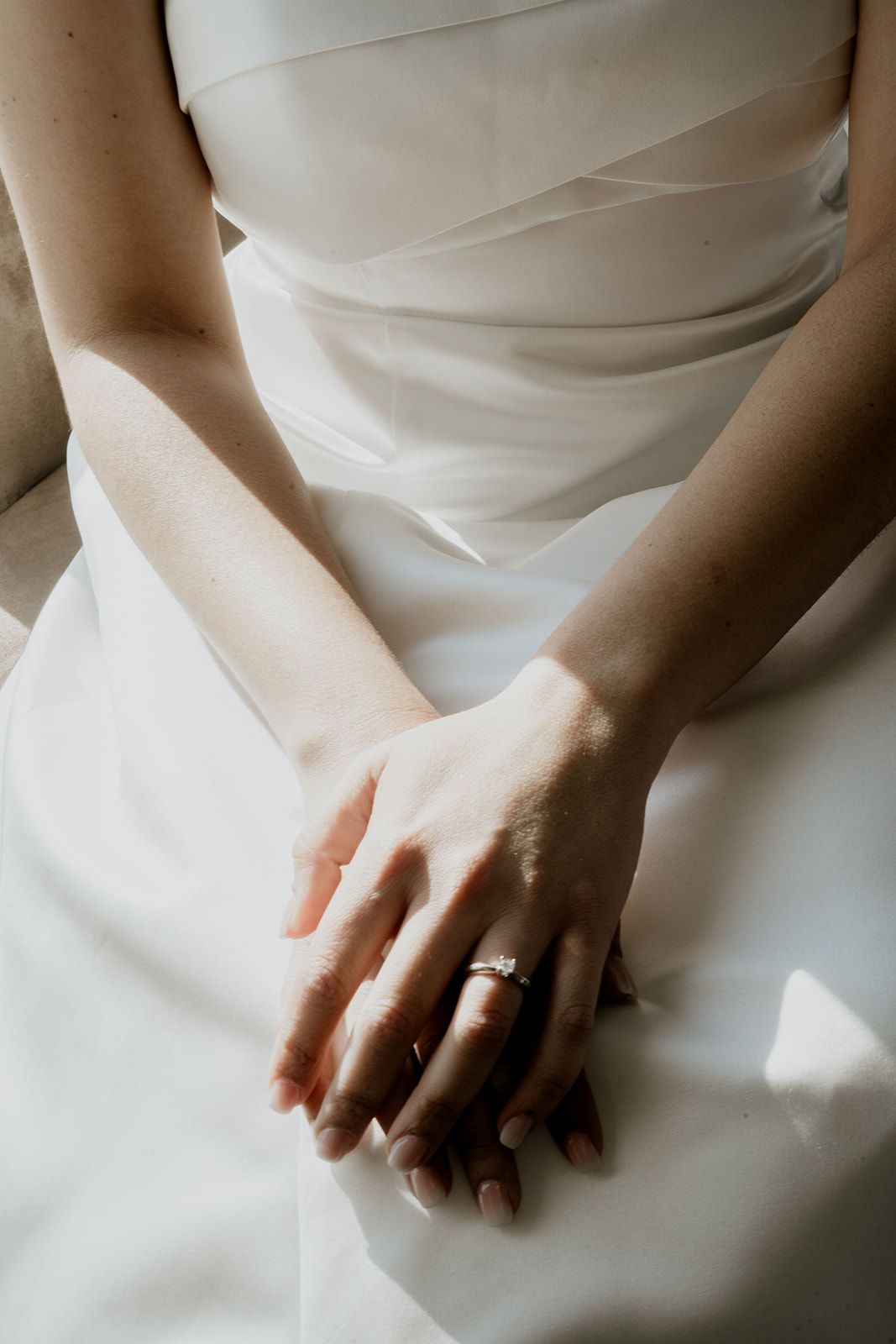 Close-up of bride's hands resting on white wedding dress with diamond engagement ring in sunlight.