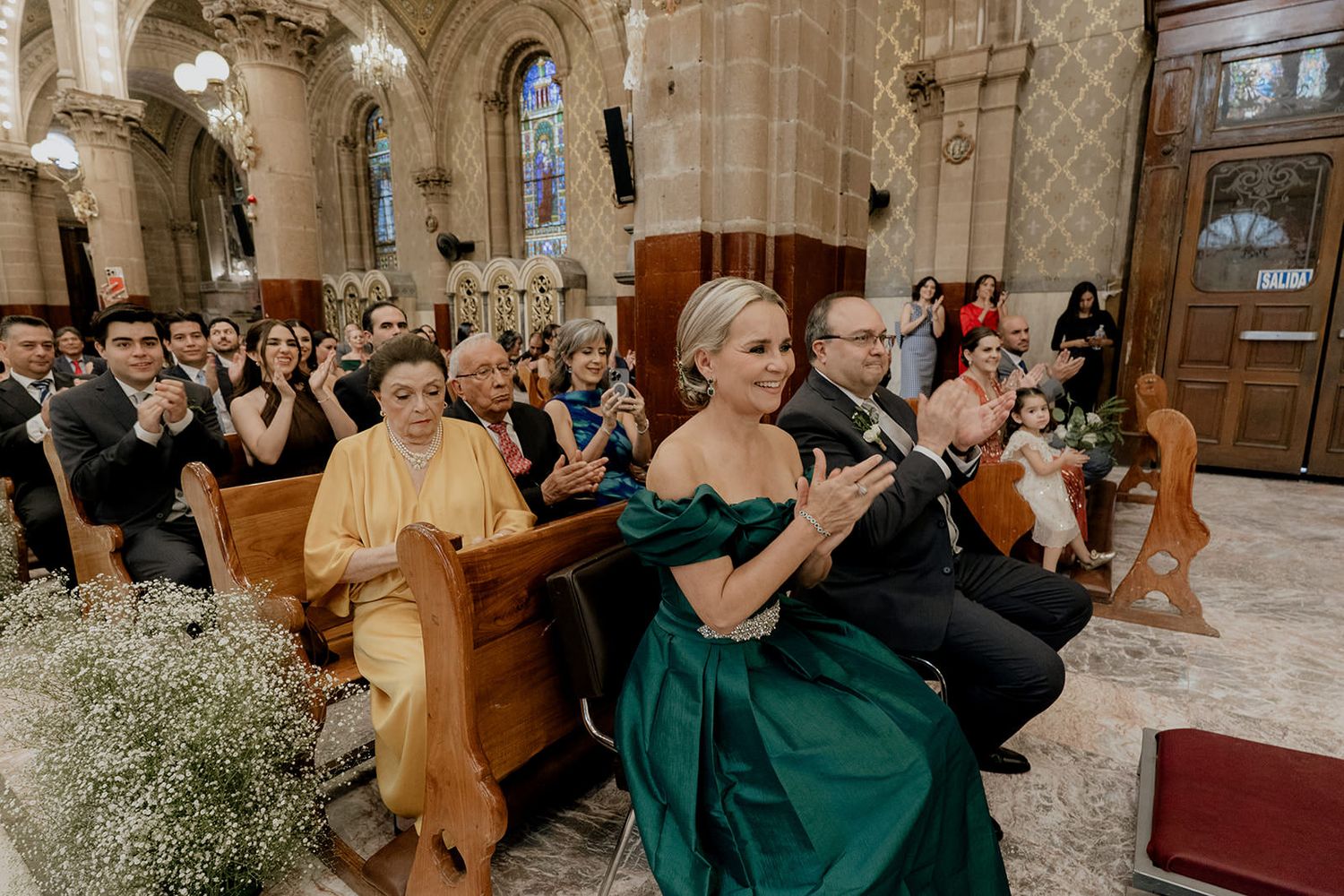 Guests in formal attire applaud during a wedding ceremony inside an ornate church with stone architecture.