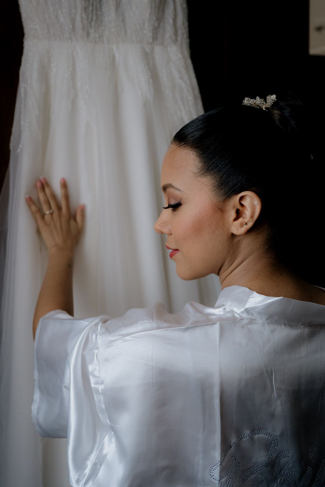Bride in white satin robe admiring her wedding dress, hair up with crystal accessory, elegant bridal preparation moment.