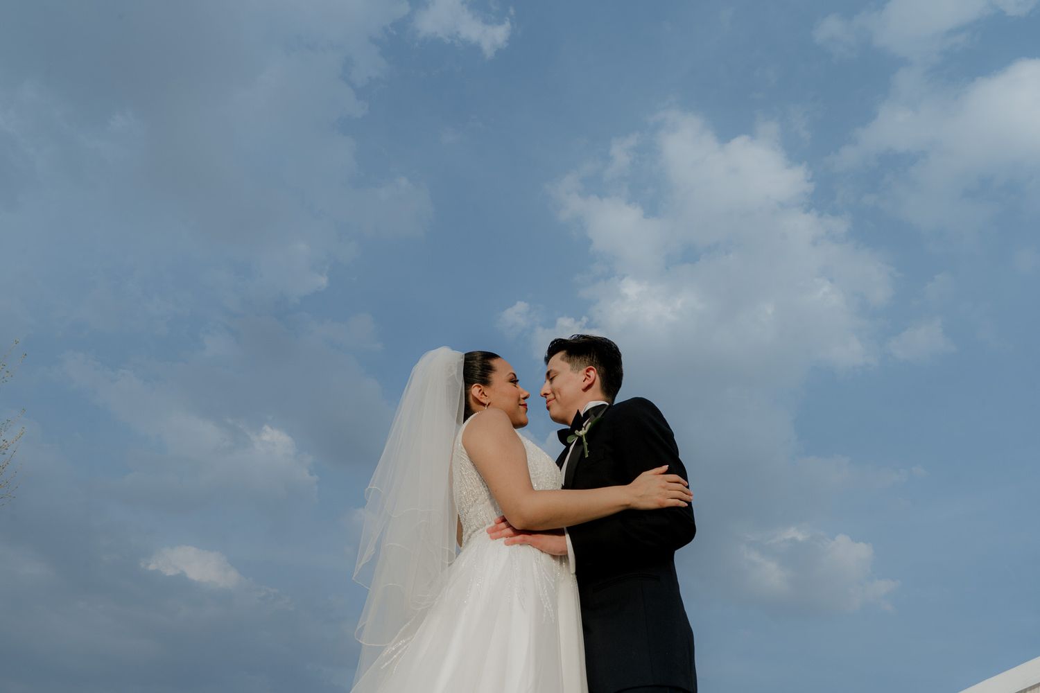 A bride and groom share a romantic moment under a blue sky with white clouds on their wedding day.