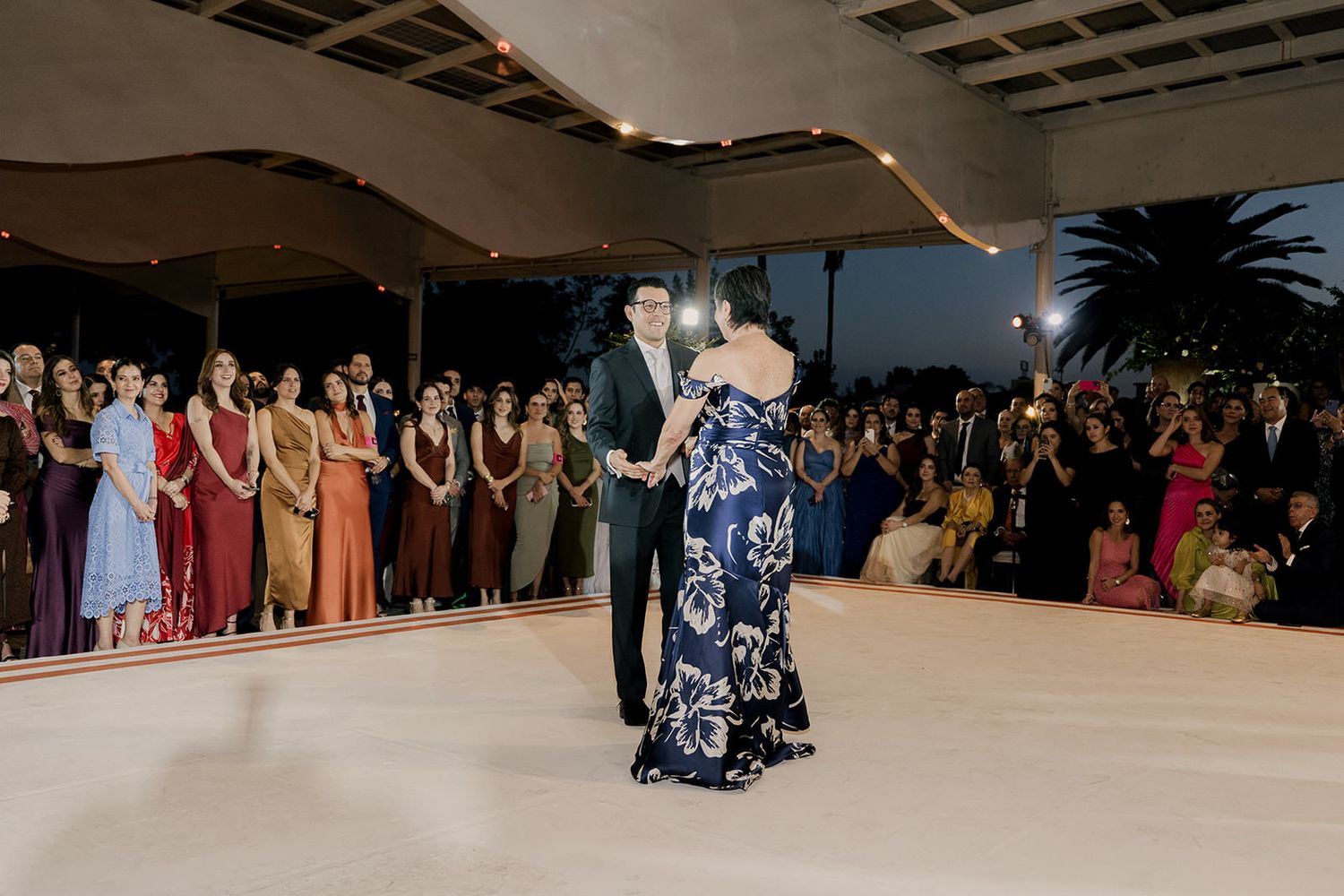 Groom dances with his mother in a navy floral gown on a white dance floor surrounded by wedding guests.