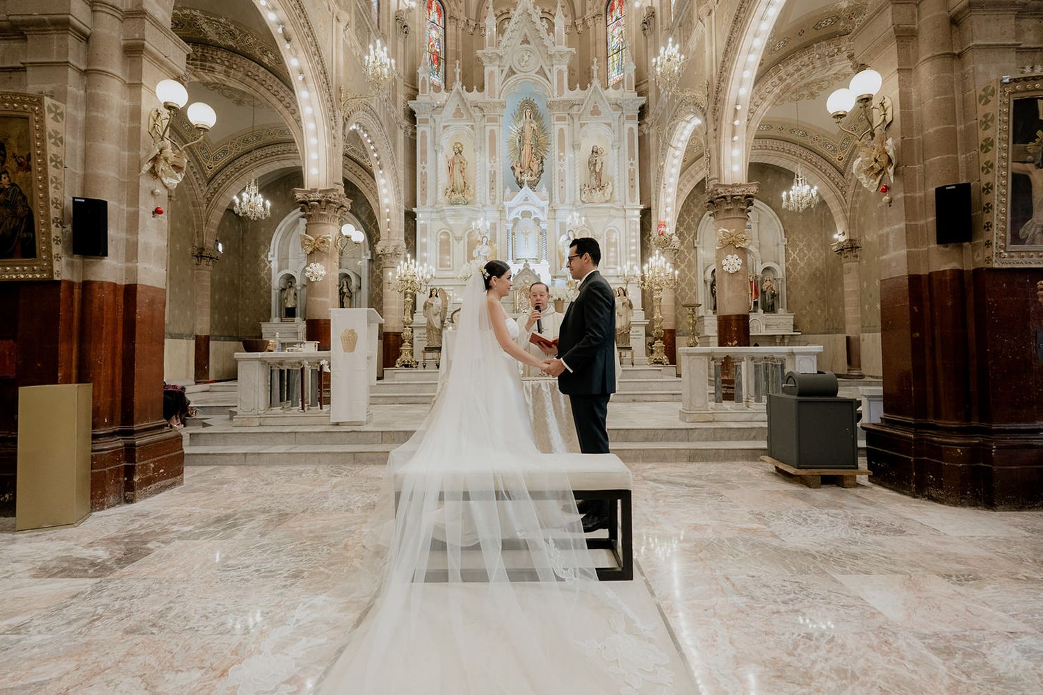 Bride and groom exchanging vows in an ornate Catholic church with a long flowing veil trailing behind.