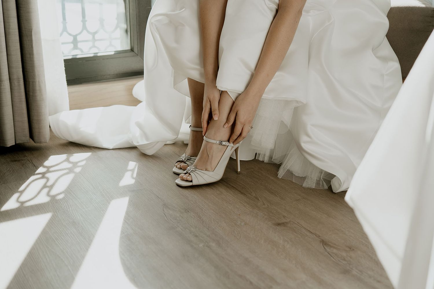 Bride in white wedding dress fastening strappy white heeled sandals in a sunlit room on wedding day.