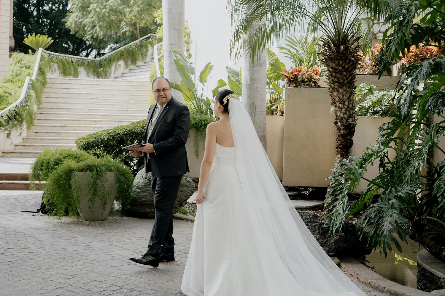 Father escorts bride in a flowing white gown and veil through a tropical garden setting.