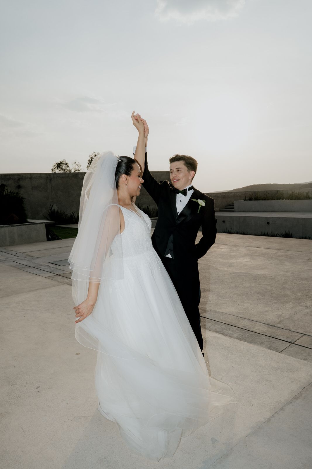 Bride and groom dancing outdoors at sunset, wearing white ball gown and black tuxedo.