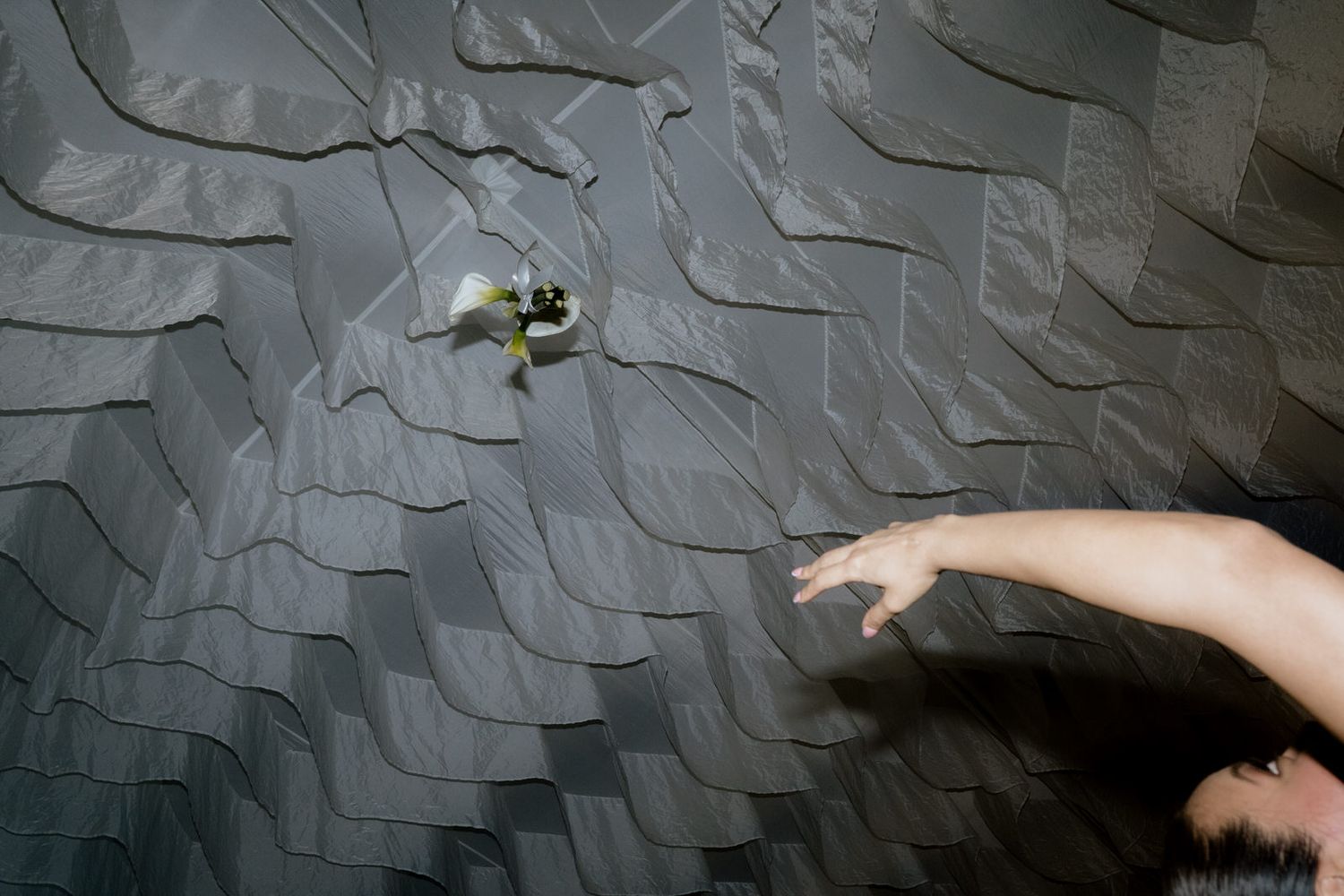 A hand reaching toward a small flower on a textured gray ruffled fabric wall.