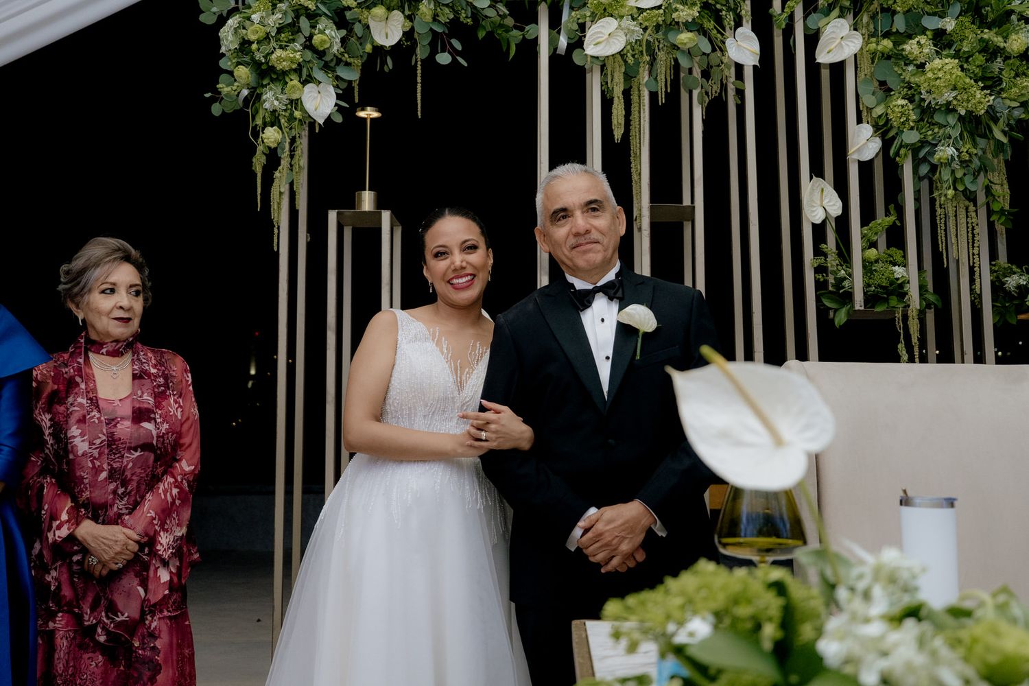 Bride in white gown stands with family at elegant wedding ceremony adorned with green floral arrangements and white calla lilies.