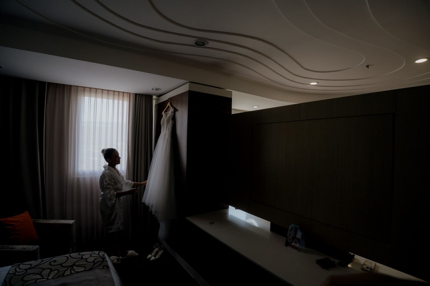 Bride holding wedding dress in dimly lit hotel room before ceremony.