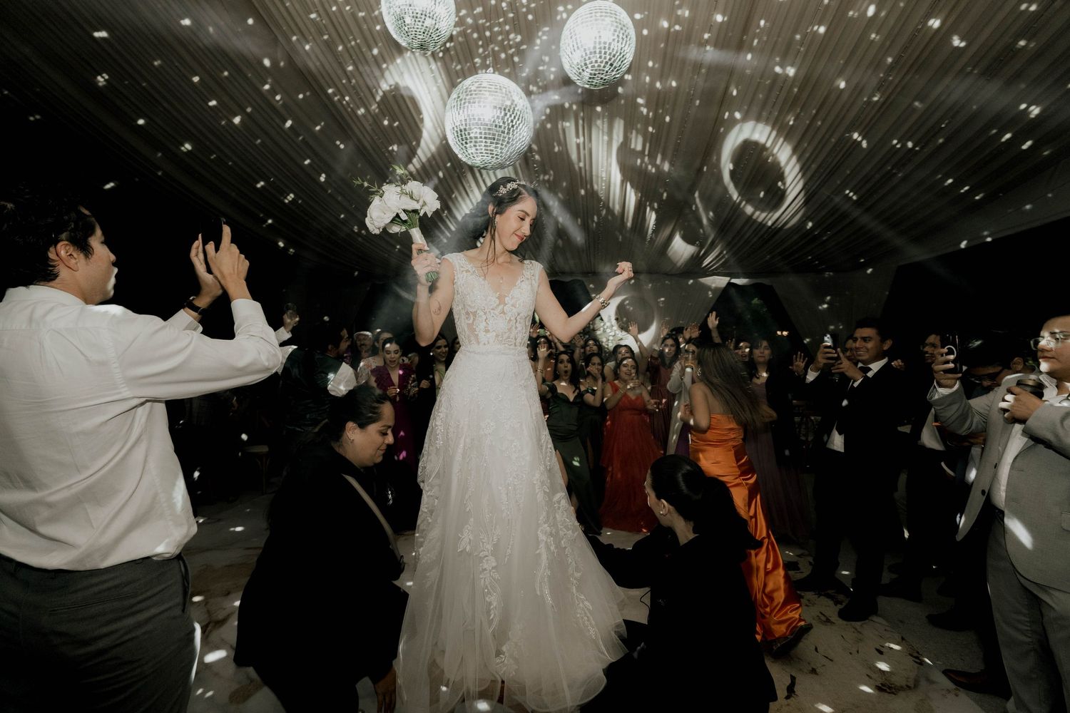 Disco balls and string lights illuminate wedding reception dance floor filled with celebrating guests.