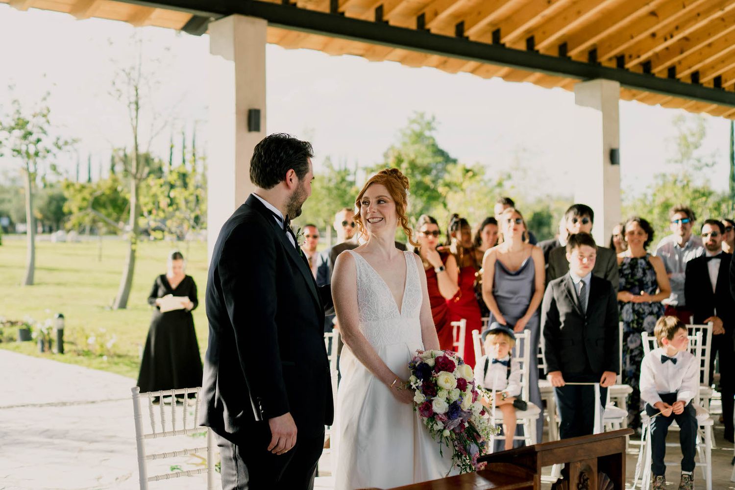 Wedding ceremony proceedings under rustic wooden pavilion with gathered guests and officiant.