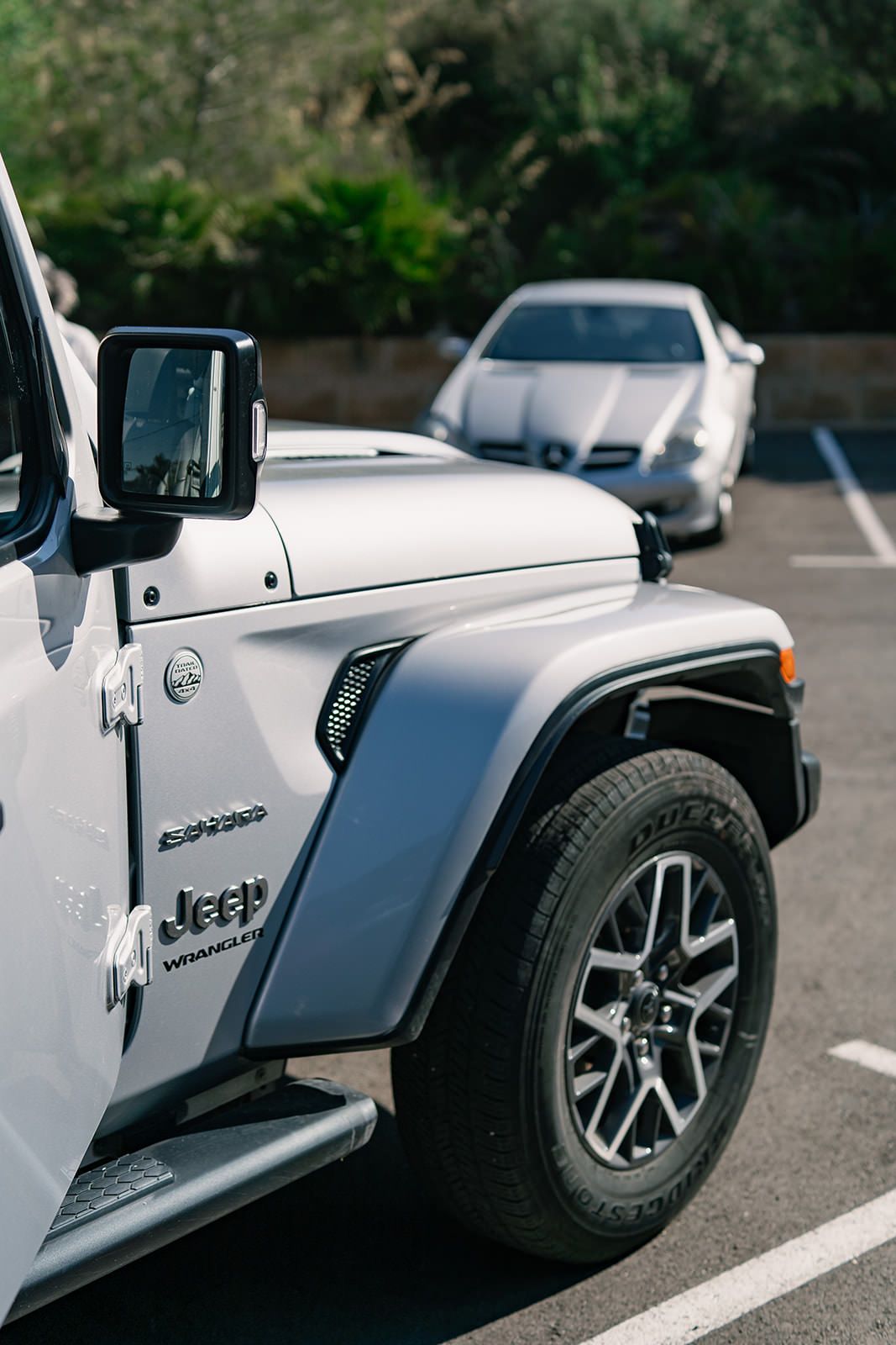Close-up view of a white Jeep Wrangler fender and wheel with custom rims parked in a lot with another car in background.