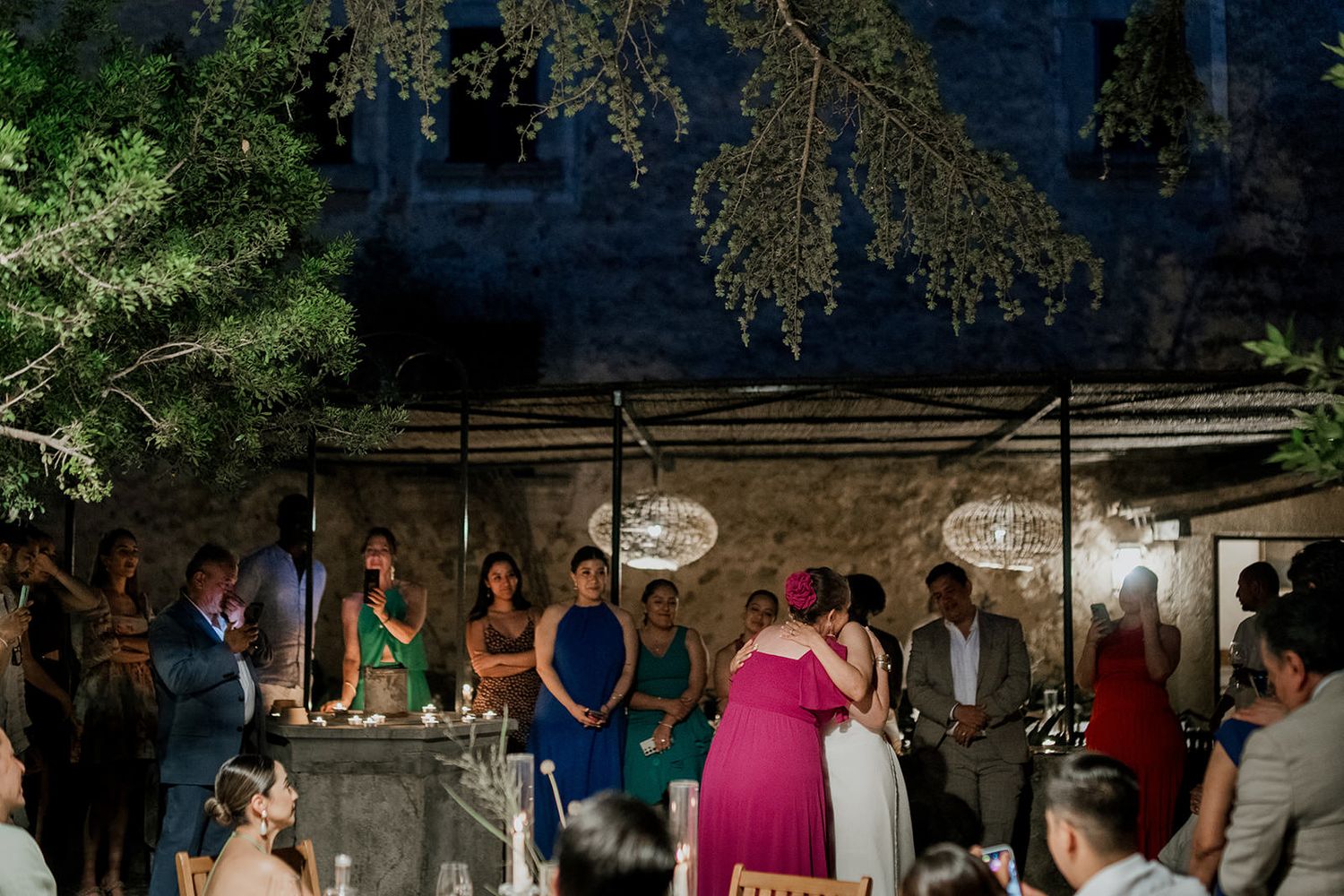 A group of people dance together at an outdoor nighttime event under trees and string lights.