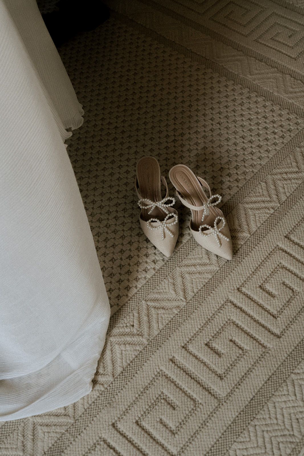 Elegant white bridal shoes with delicate bow details rest on a patterned carpet near white fabric.