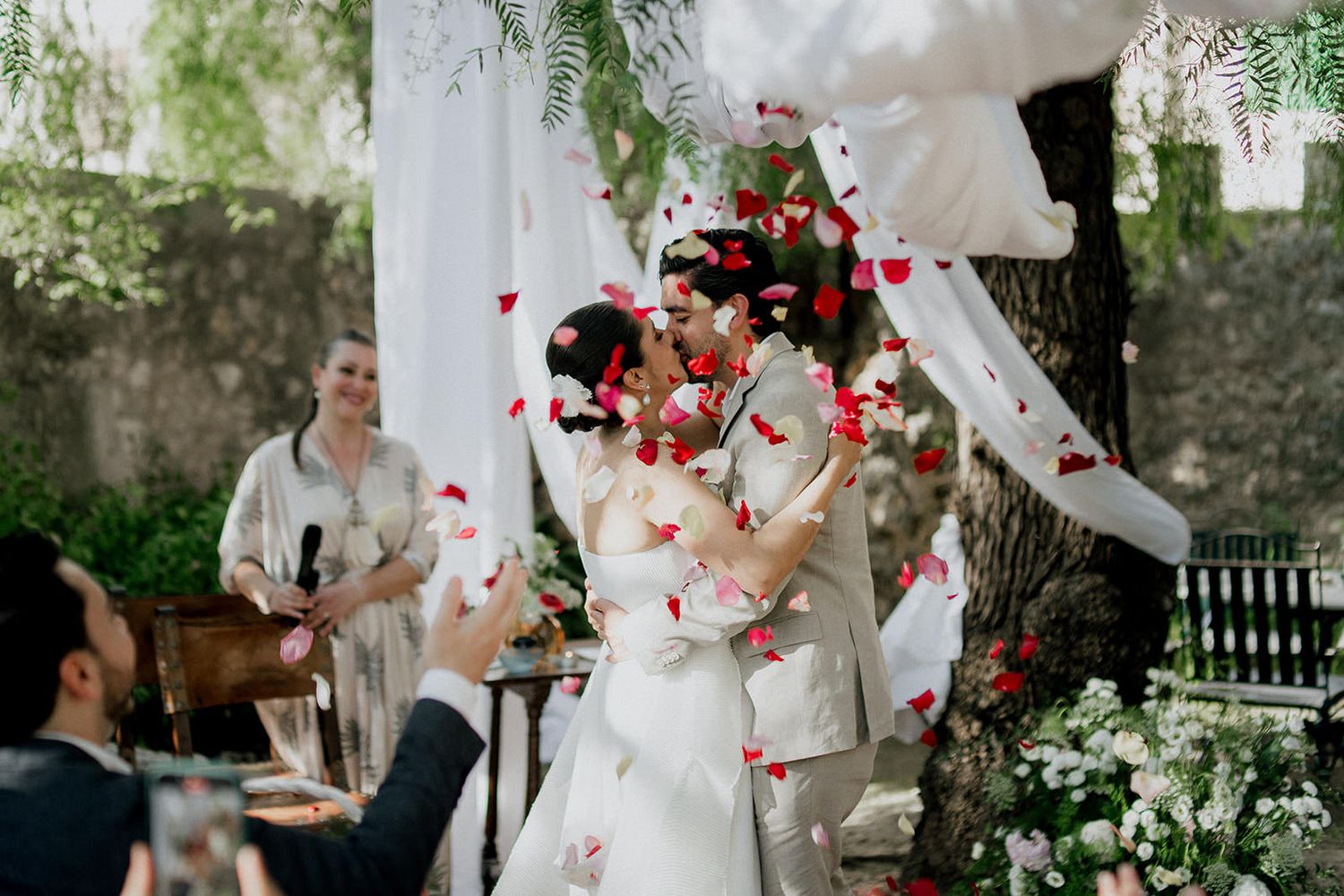 Couple in white wedding attire embraces under a tree while red rose petals fall around them in a romantic outdoor setting.