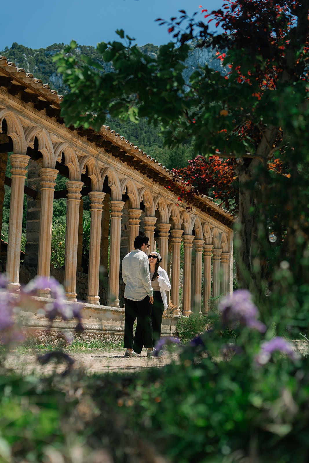 Historic stone colonnade with arched columns surrounded by blooming purple flowers and lush greenery.