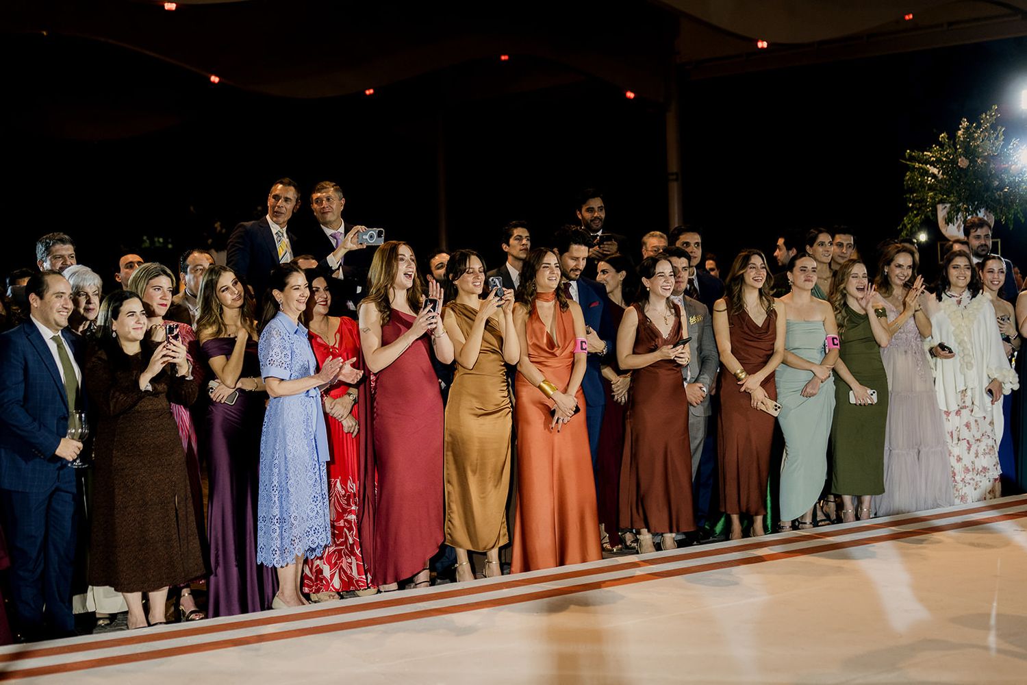 Elegantly dressed wedding guests in colorful gowns applauding on a dance floor at a formal reception.