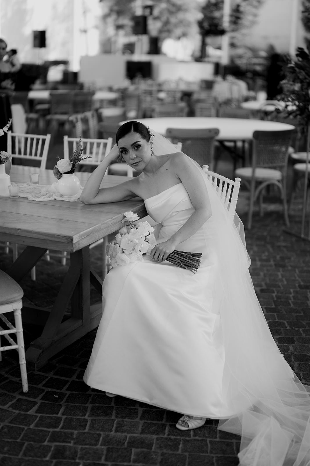Bride in white strapless gown sits at wooden table holding bouquet, black and white wedding photo.