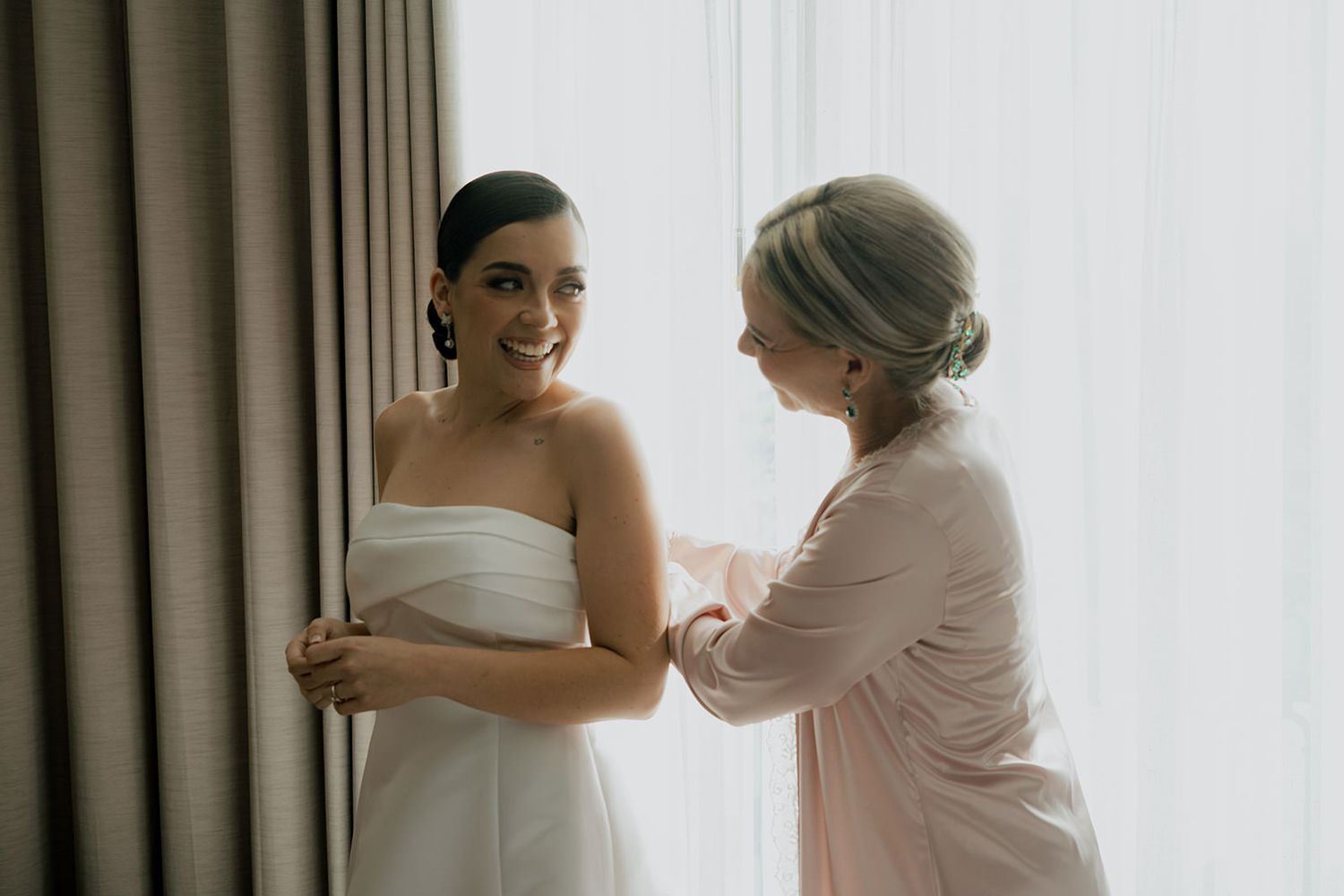 Bride in white strapless gown being helped by bridesmaid in pink dress near curtained window.