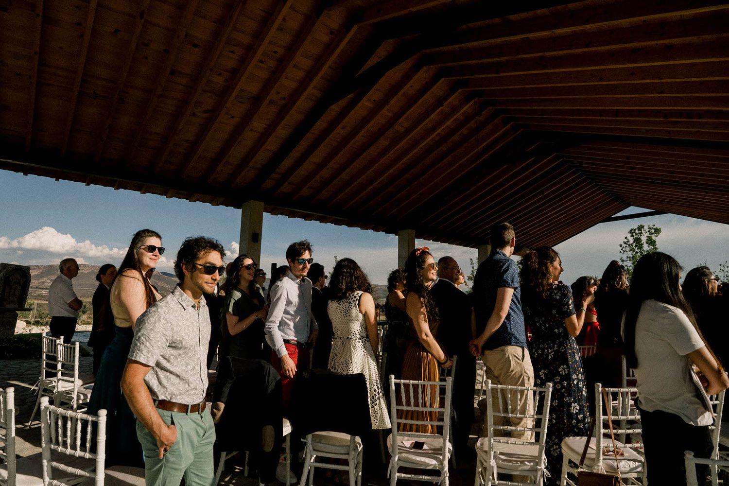 Wedding guests seated under rustic wooden beamed pavilion during outdoor ceremony.