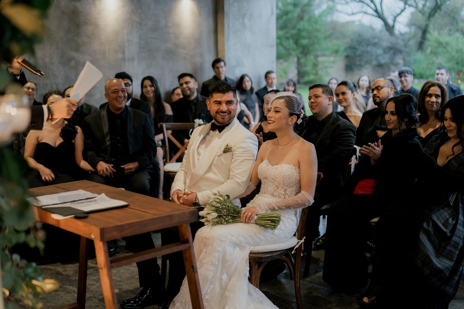 A couple sits at a wooden table during their wedding ceremony surrounded by guests in formal attire.