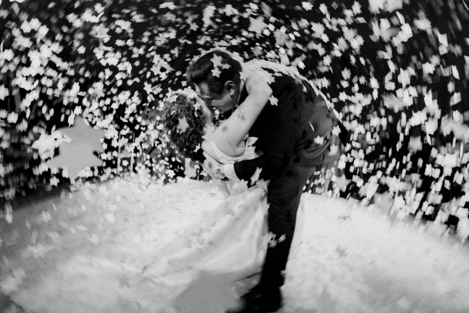 A romantic black and white photo of a couple dancing in a swirl of confetti at their wedding celebration.