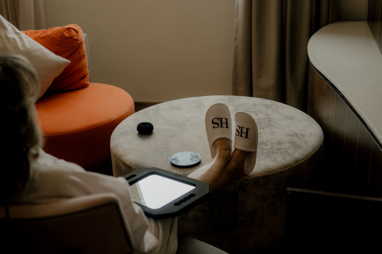 Hotel slippers with SH monogram on a round table beside an orange chair in a modern hotel room.