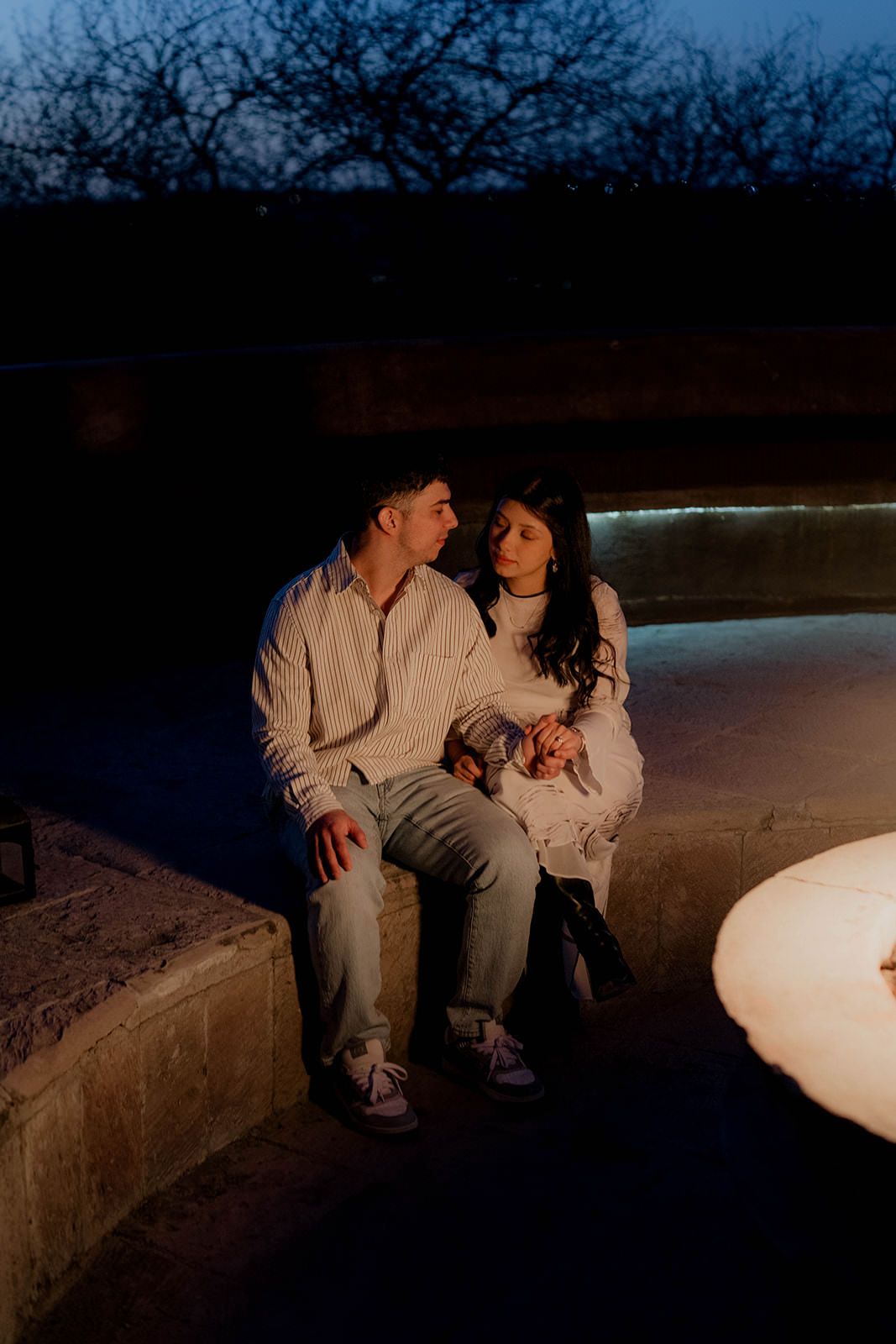 Couple sitting together on concrete bench by glowing fire pit at night.