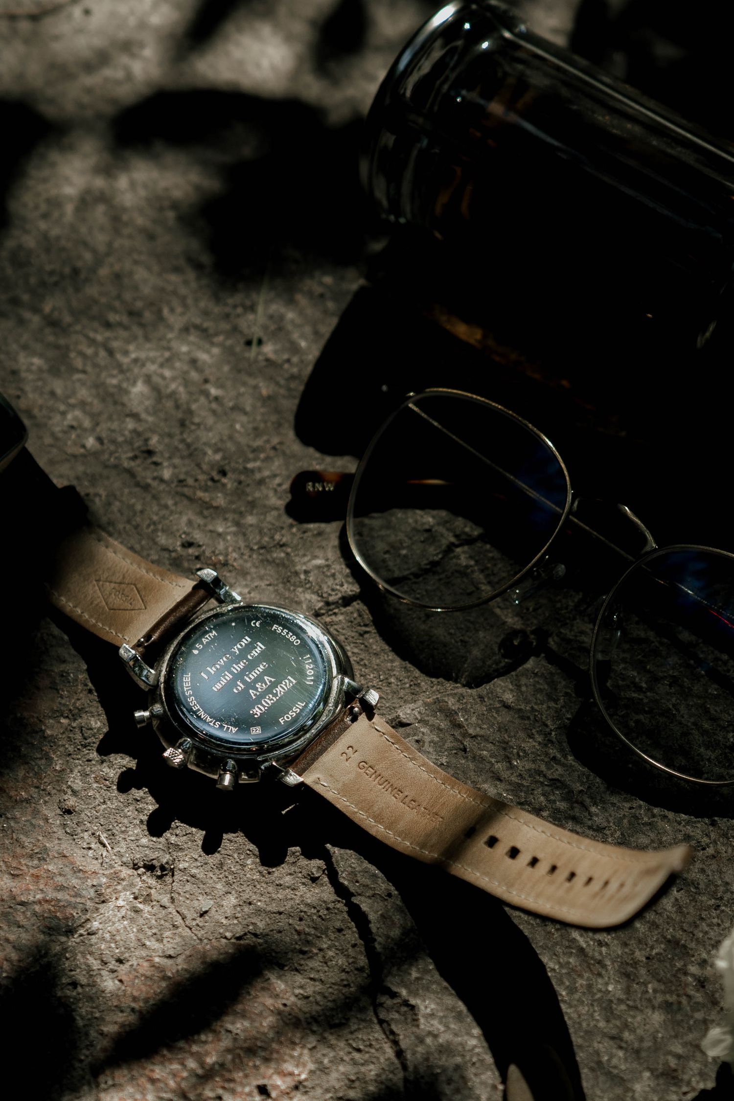 Modern wristwatch with leather strap and sunglasses on textured surface in moody lighting.