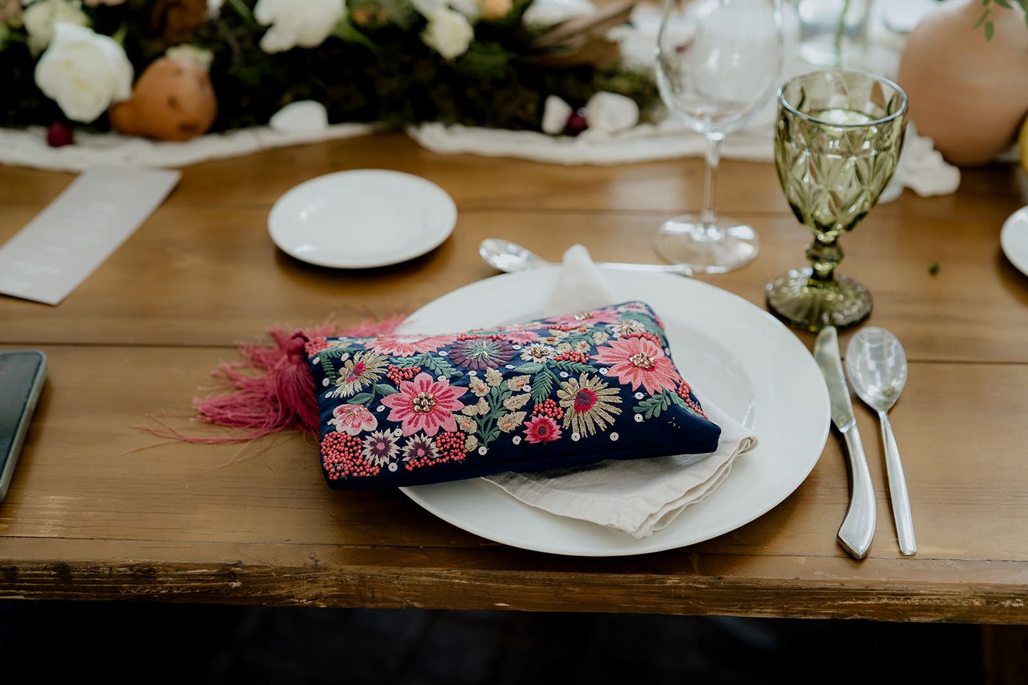 Elegant wedding place setting with floral embroidered navy pillow on white plate, gold cutlery on rustic wood table.
