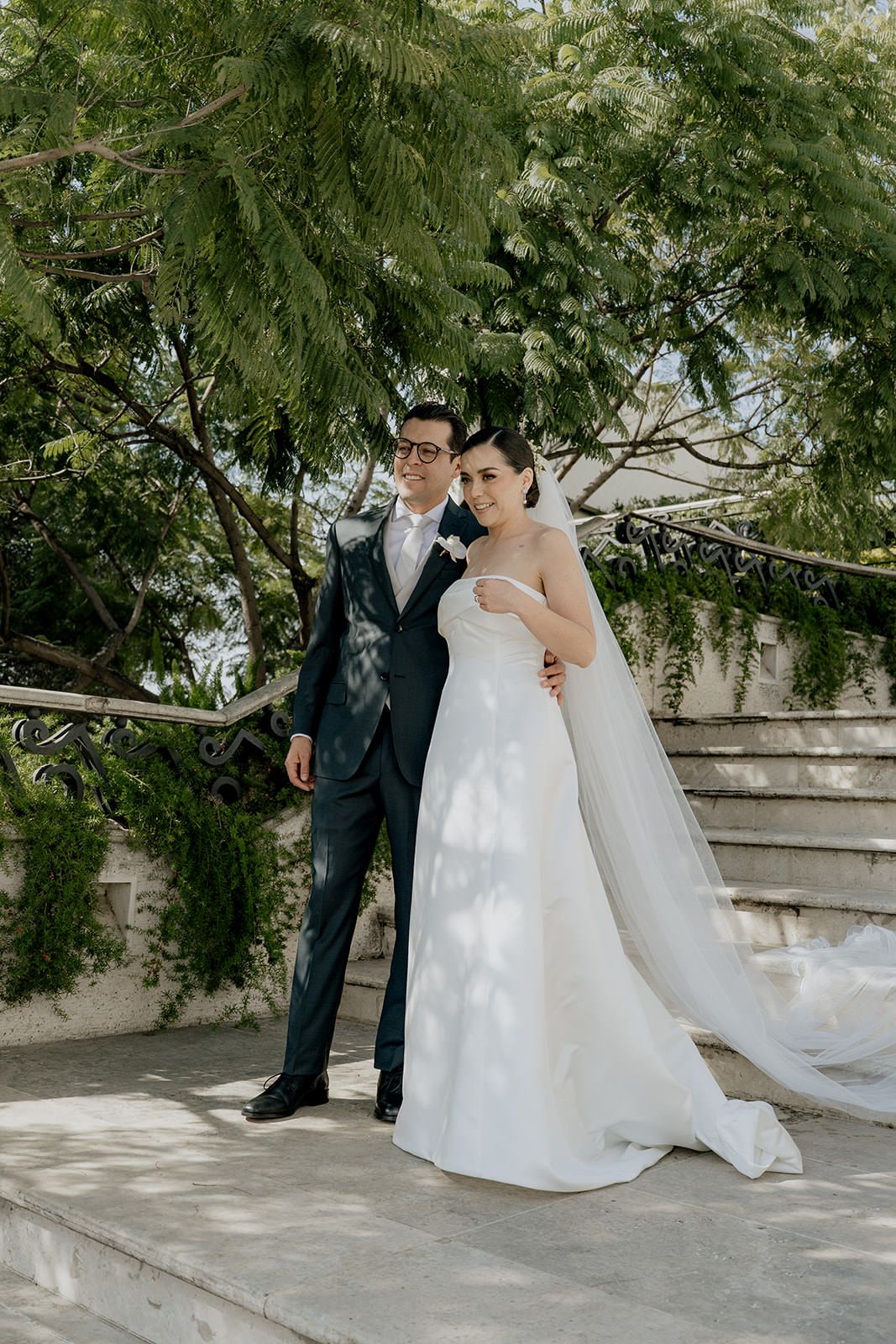 Elegant bride and groom posing together outdoors under lush green trees on their wedding day.