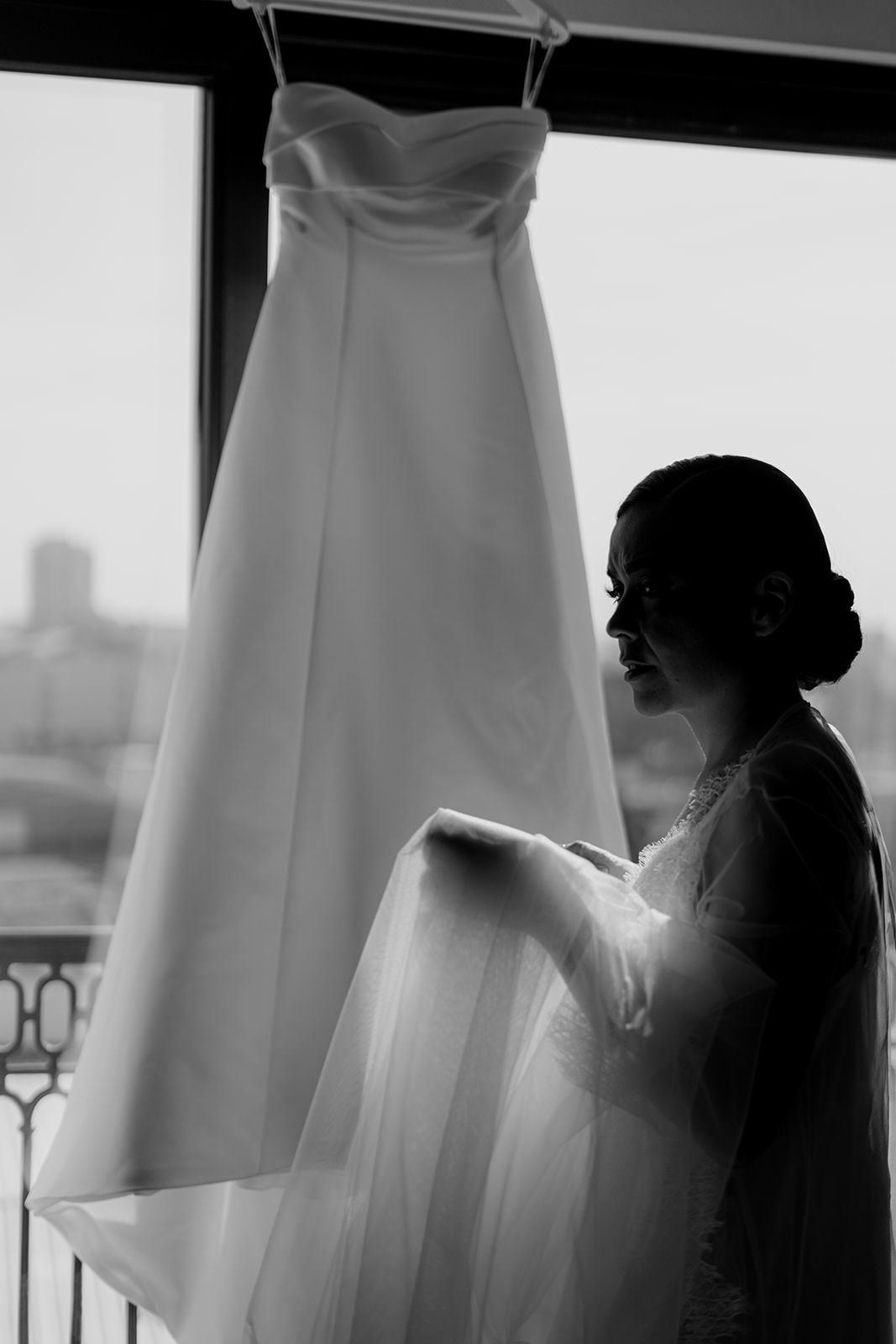 Bride in black and white photo touching her wedding dress hanging by a window.